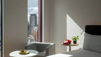 Habitación de hotel con un ventanal grande, una mesa redonda con un plato de fruta, una silla, una mesita de noche con una planta y un teléfono rojo, y una cama con cabecera negra.