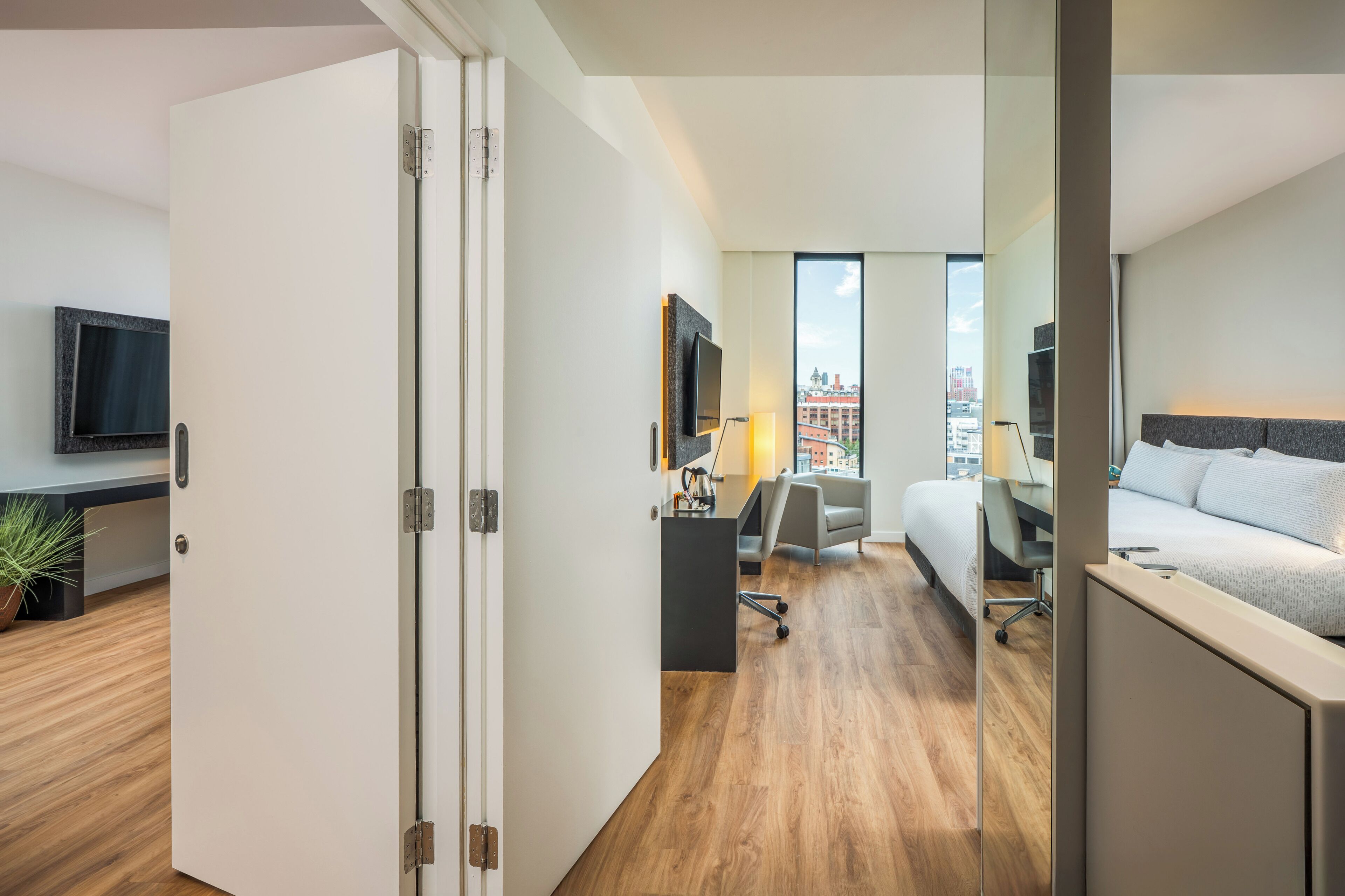 A modern hotel room with a wooden floor, a bed, a desk with a chair, a TV, and a city view.