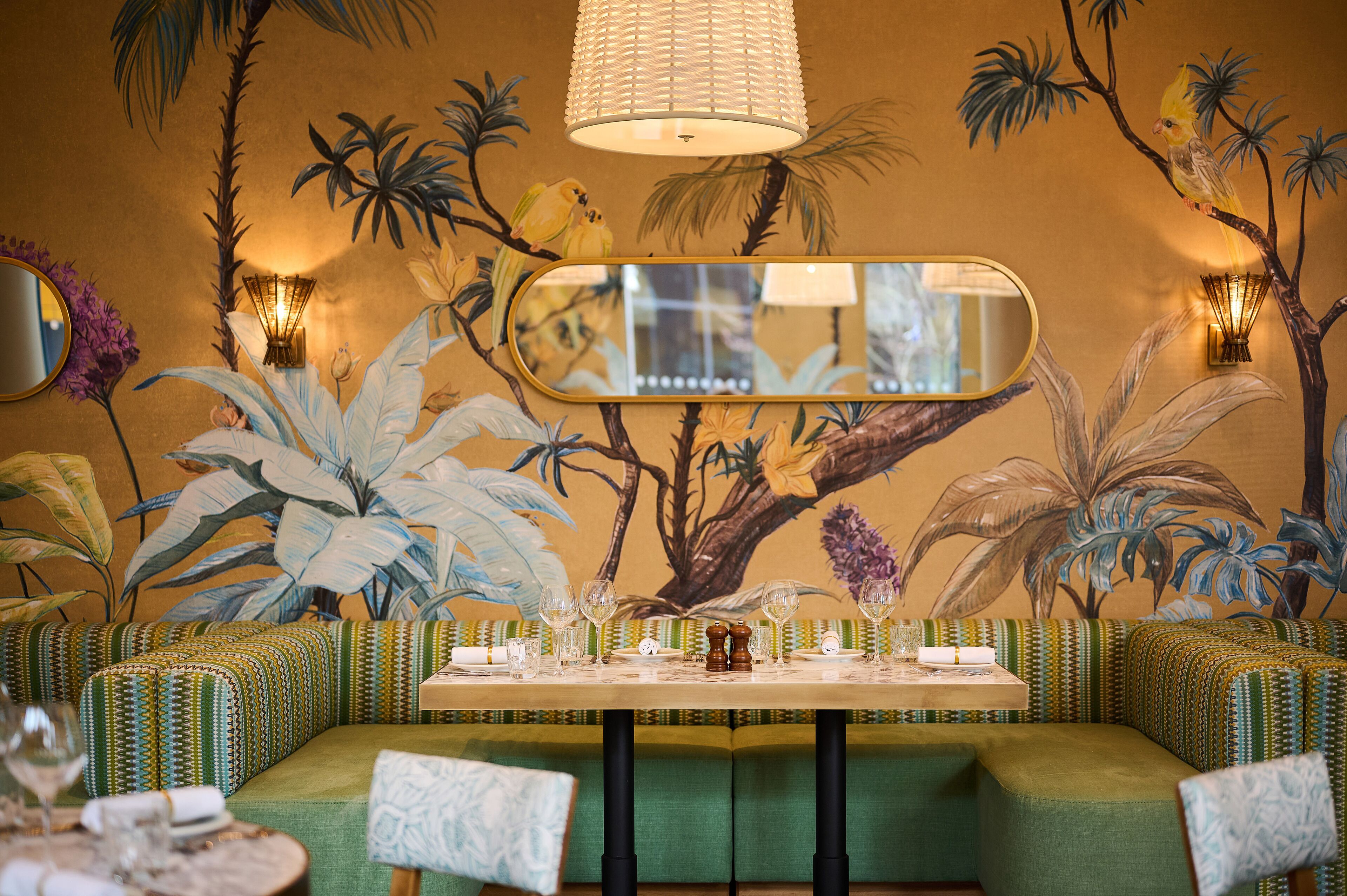 A dining area with a mural of tropical plants, a long table set for dining, and green upholstered seating.