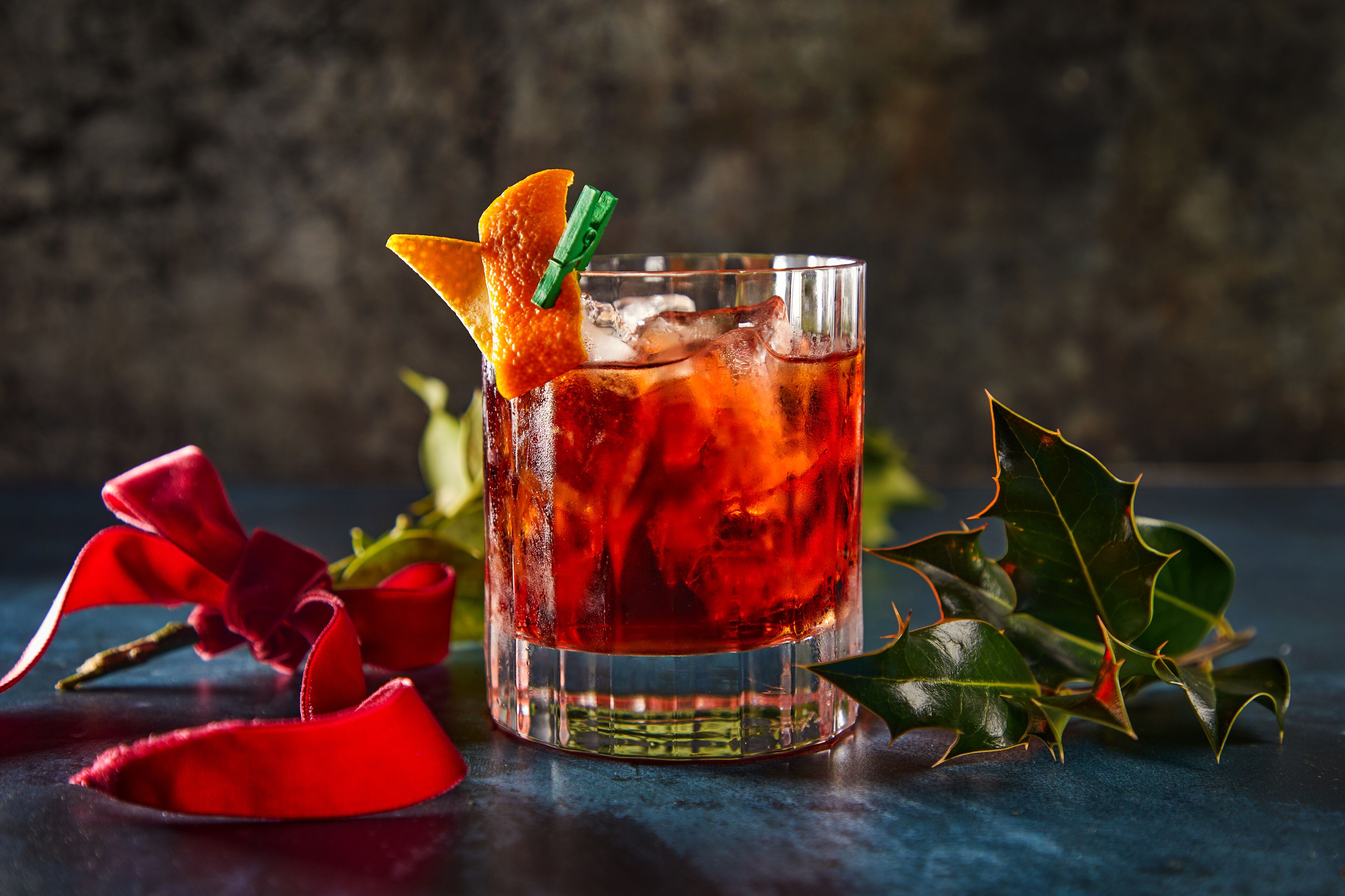 A glass of red beverage with ice cubes, garnished with an orange slice and a green leaf, on a dark surface with a red ribbon and green foliage.