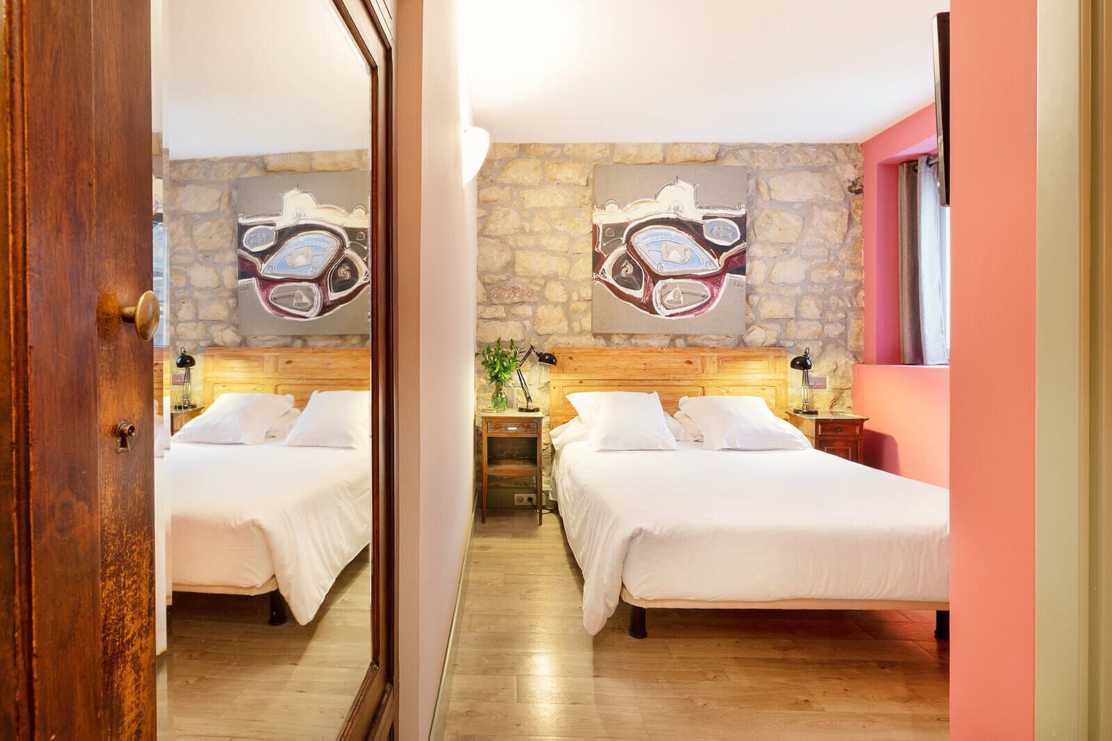 A hotel room with two beds, a stone wall, and a mirror reflecting the room.