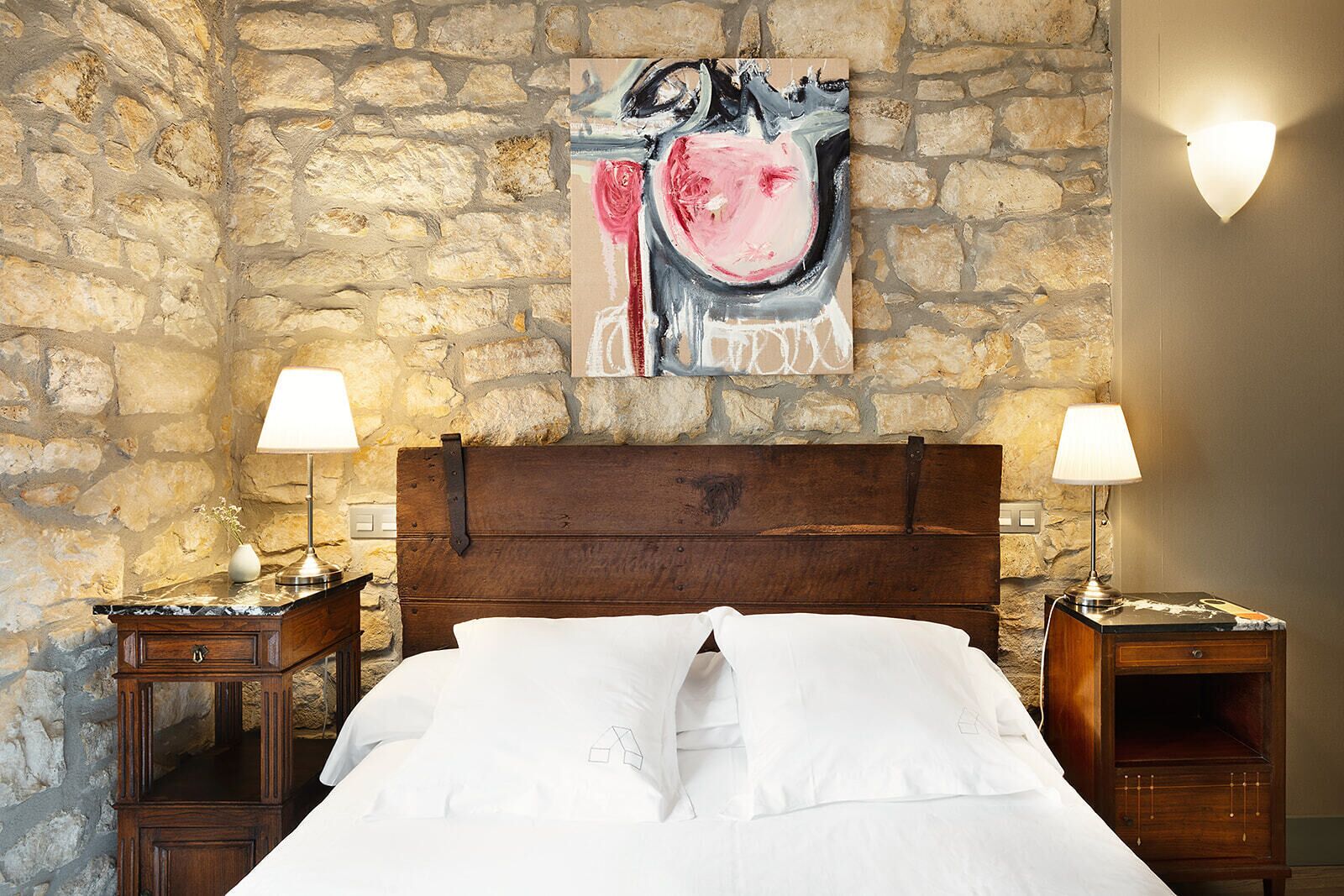 A bedroom with a stone wall, a bed with white linens, two bedside tables with lamps, and a painting above the bed.