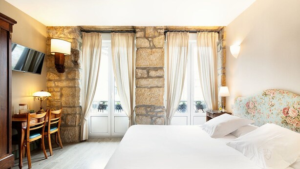 A hotel room with a large bed, a desk with a chair, a TV, and stone walls. - Okako Hotel (San Sebastián)