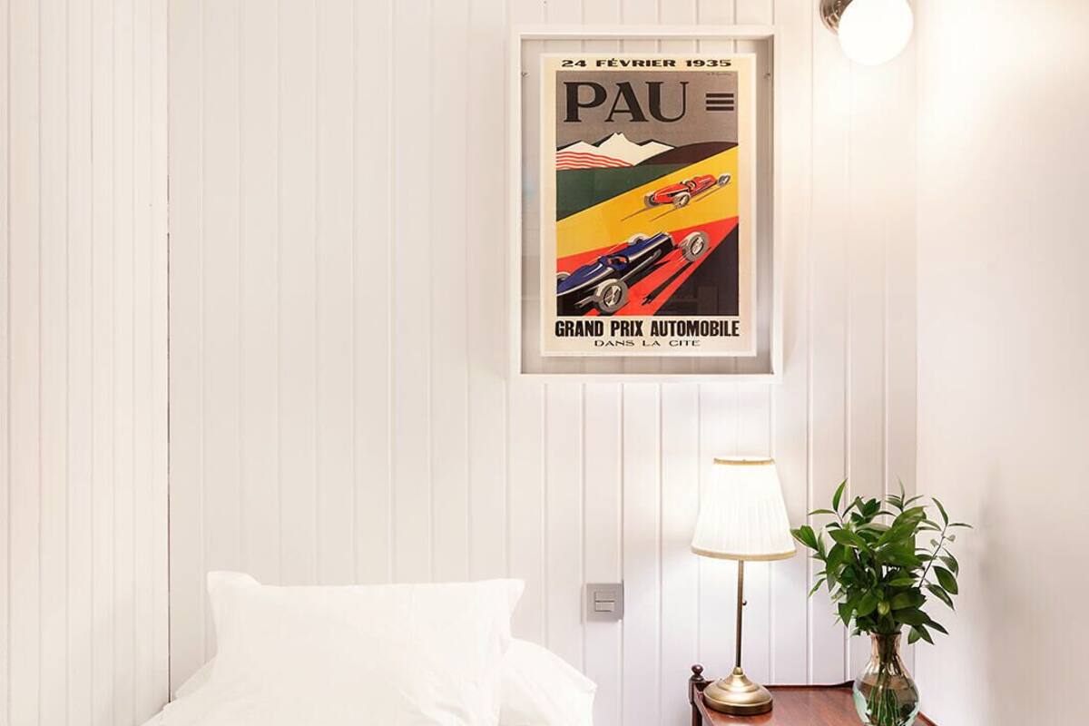 A hotel room with a bed, a nightstand with a lamp, a framed poster on the wall, and a potted plant.