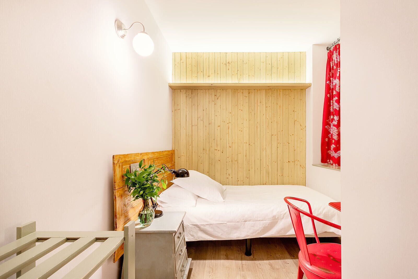 Un dormitorio pequeño y ordenado con cabecera de madera, mesita de noche con lámpara, una silla roja y cortina roja.