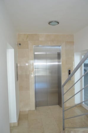 A modern elevator with a metallic finish, located in a hallway with beige tiled walls and floor. - Apartment Silvestre (Santo Domingo)