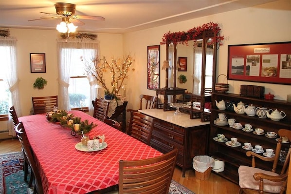A dining room with a red checkered tablecloth, wooden chairs, a chandelier, a mirror, and a shelf with teapots.