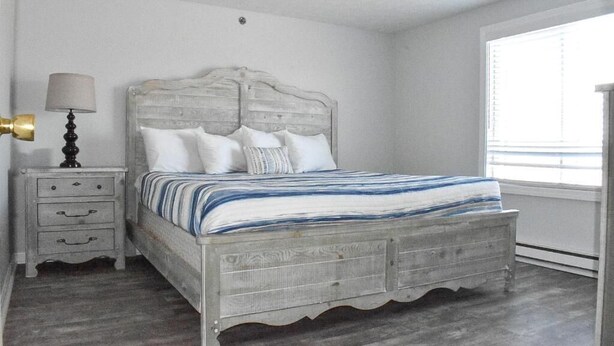 A bedroom with a wooden bed, a nightstand with drawers, a lamp, and a window with blinds. - Grand Beach Inn (Old Orchard Beach)