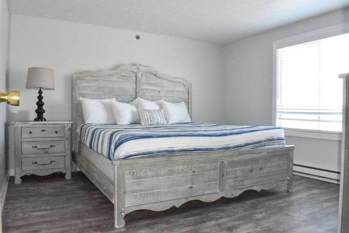 A bedroom with a wooden bed, a nightstand with drawers, a lamp, and a window with blinds.