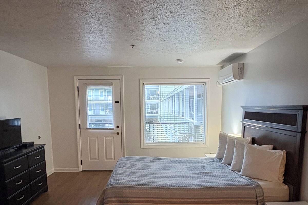 A bedroom with a bed, dresser, and a TV. There are two windows with blinds, one of which has a view of a building.