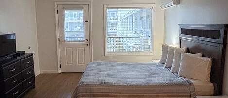 Une chambre à coucher avec un lit, une commode et un téléviseur. Il y a deux fenêtres équipées de stores, et l’une d’elles offre une vue sur un immeuble.