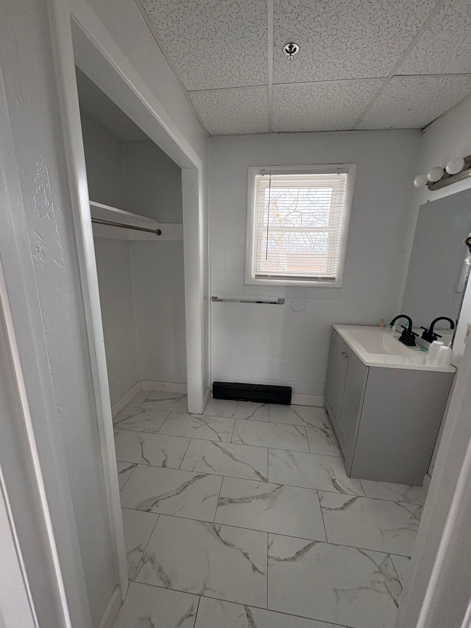 A bathroom with a white tiled floor, a window with blinds, a towel rack, a sink with a mirror, and a door leading to another room.