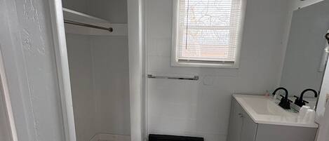 A bathroom with a white tiled floor, a window with blinds, a towel rack, a sink with a mirror, and a door leading to another room.