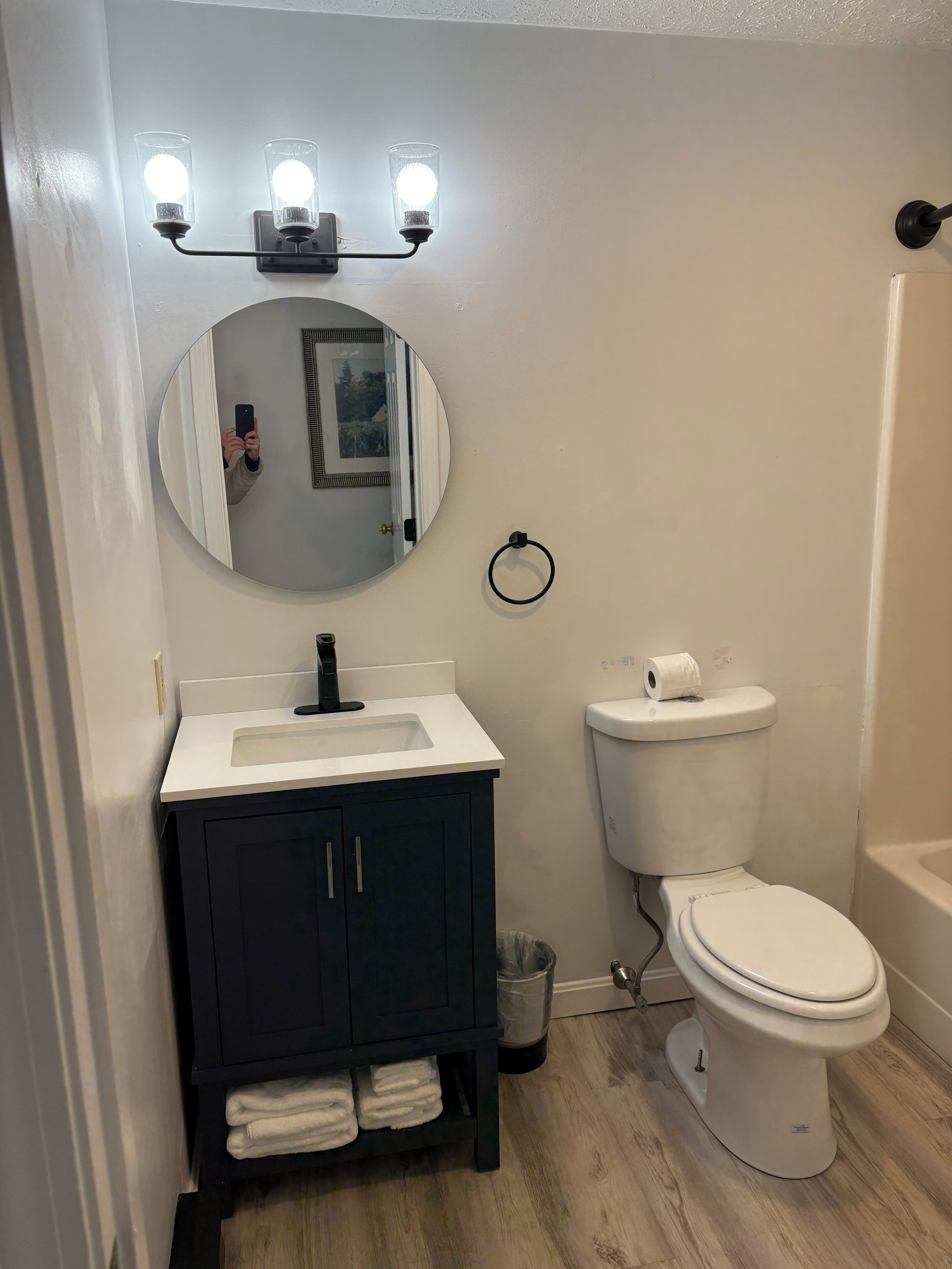 Une salle de bain avec un lavabo blanc, des meubles sombres, un miroir rond et des toilettes.