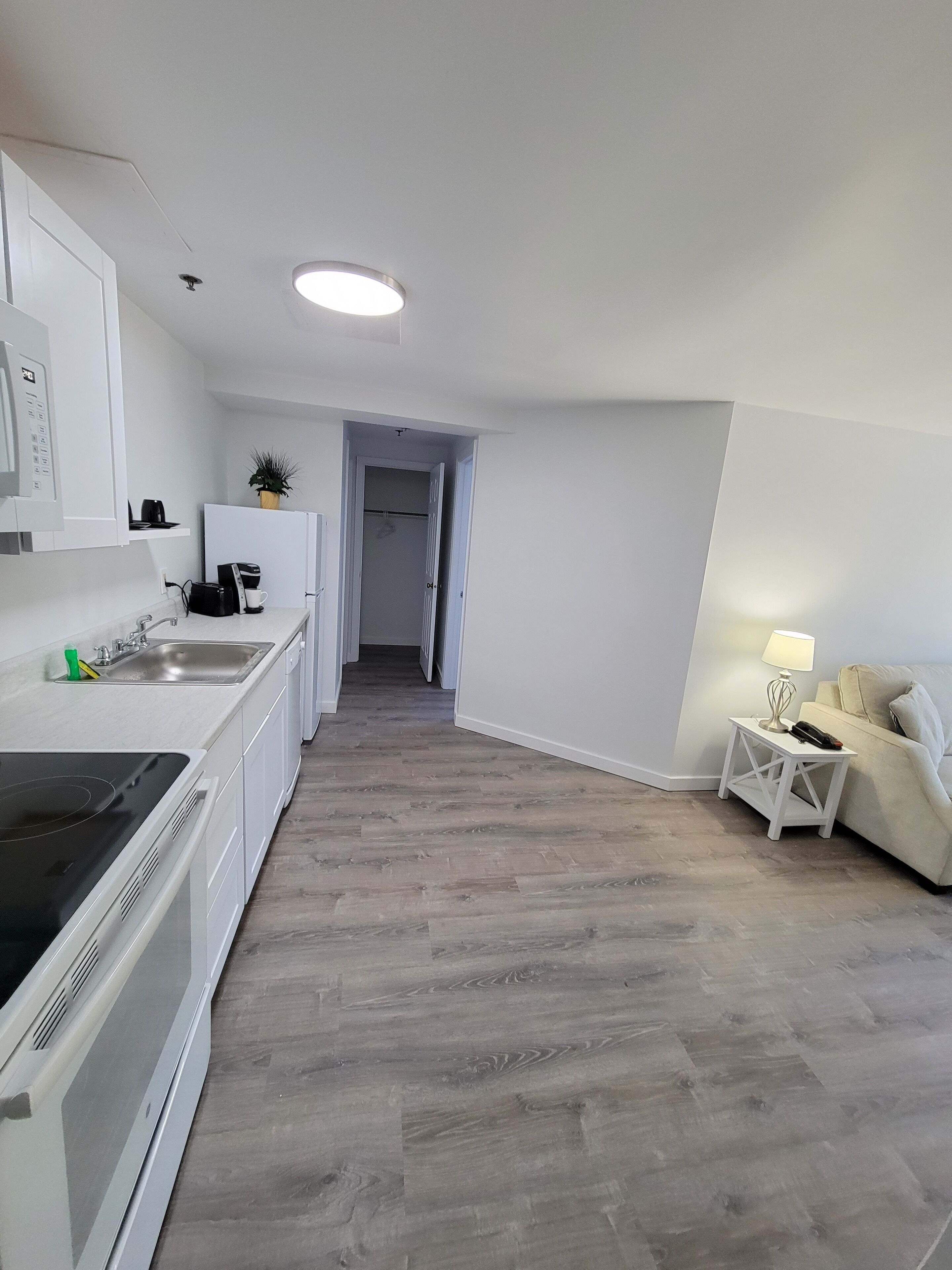 Een moderne keuken met witte kasten, een ingebouwde oven en een koffieapparaat. Een hal leidt naar een slaapkamer met een wit bed en een nachtkastje.