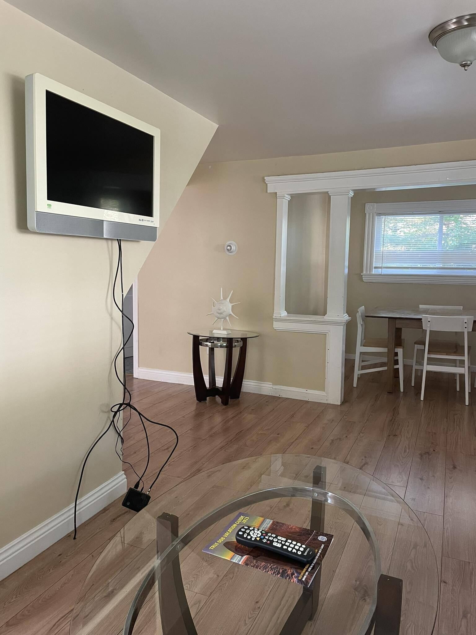 A living room with a flat-screen TV mounted on the wall, a glass coffee table with a remote control, and a dining area with white chairs and a window.