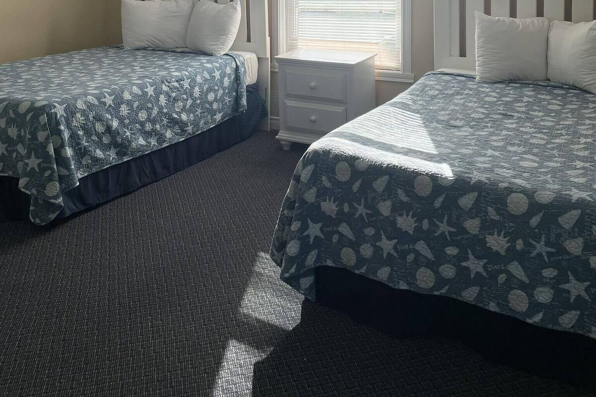 A twin bedroom with two beds, each with a white headboard and a blue bedspread with star patterns. There is a window with blinds, a white nightstand, and a ceiling light fixture.