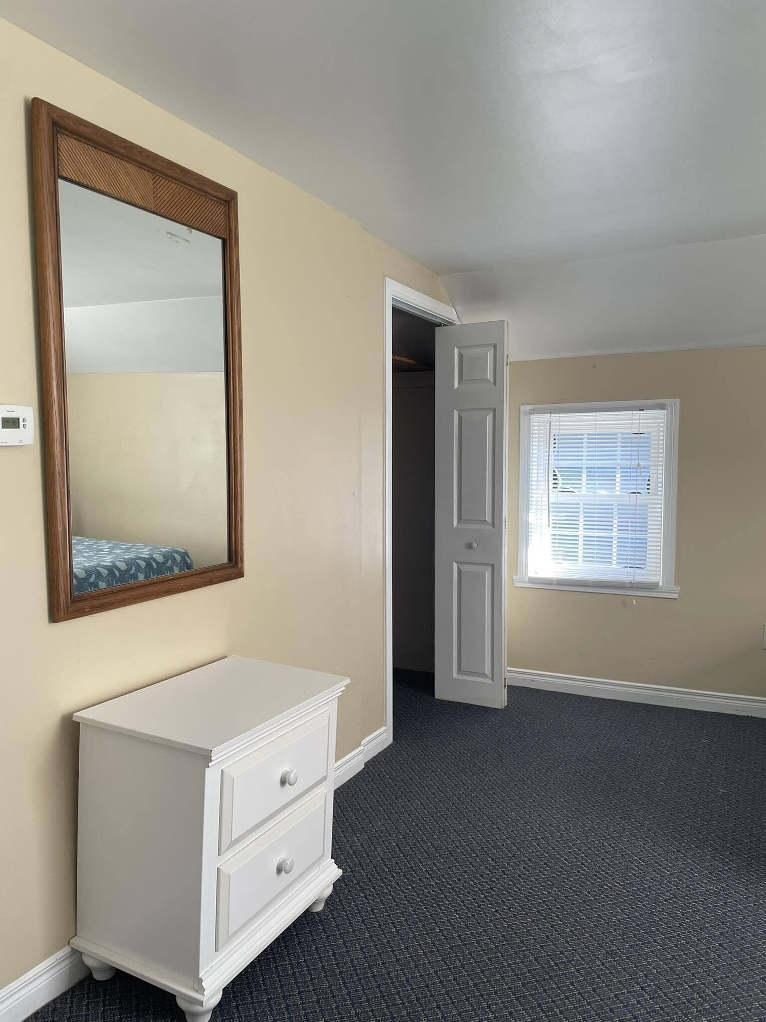 Quarto com penteadeira branca, um espelho grande, porta que dá para outro cômodo e janela com persianas.