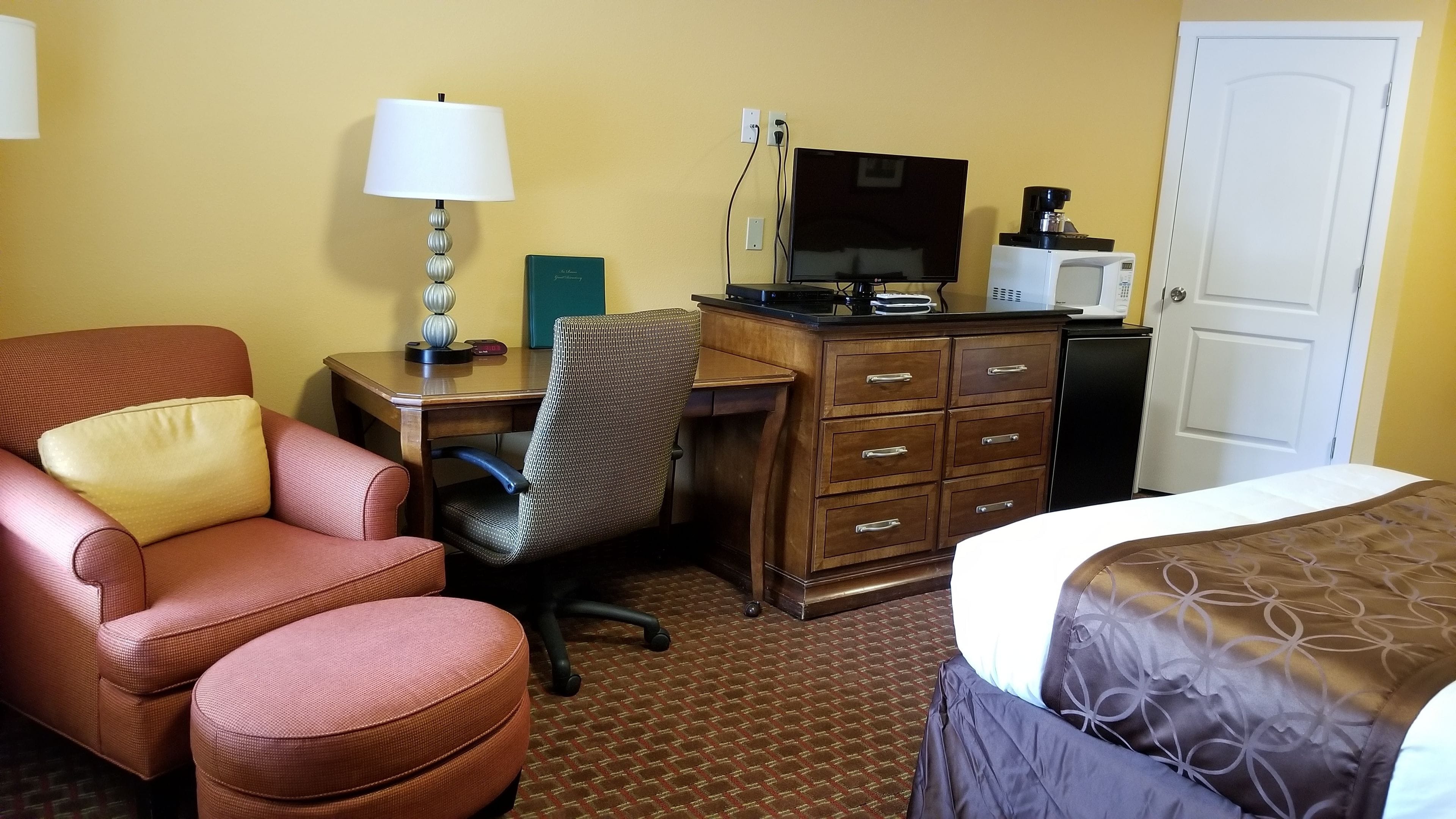 A hotel room with a desk, chair, bed, dresser, lamp, and a television.