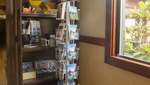 A corner with a wooden bookshelf containing various items, a white stand holding postcards, and a window with a view of greenery. - The Forks Motel (Forks)