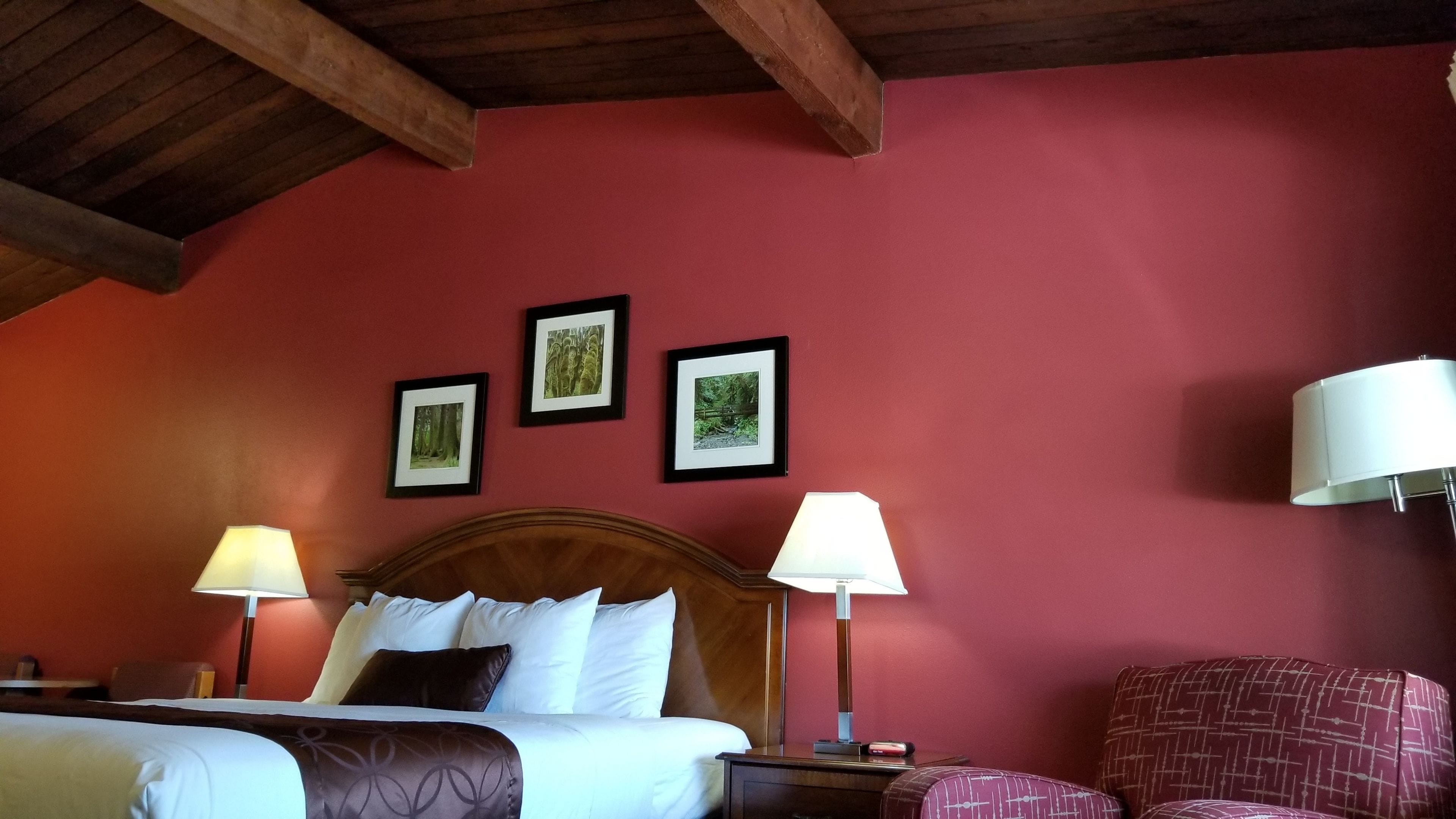 Une chambre d’hôtel avec un plafond en bois, un lit avec une literie blanche, deux lampes de chevet, une chaise et trois tableaux encadrés accrochés au mur.