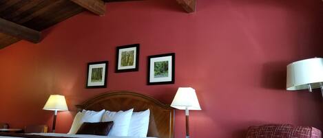 Une chambre d’hôtel avec un plafond en bois, un lit avec une literie blanche, deux lampes de chevet, une chaise et trois tableaux encadrés accrochés au mur.