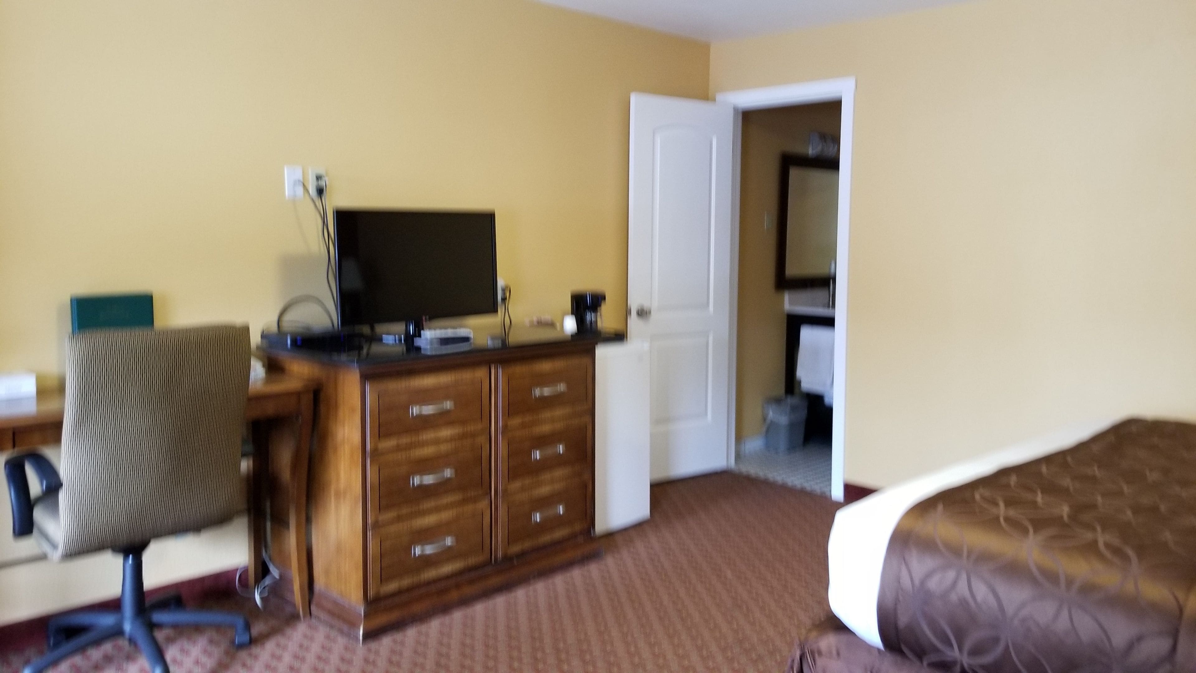 A hotel room with a wooden desk, a chair, a television, a dresser, and a bed.