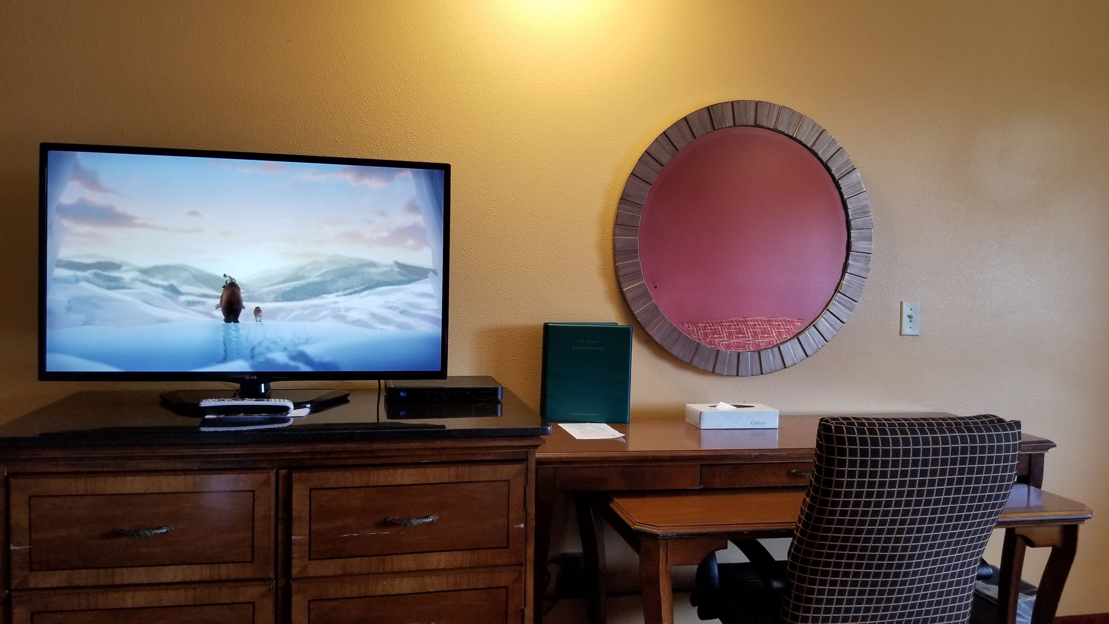 Une chambre d’hôtel avec un téléviseur diffusant un paysage enneigé, un miroir rond, un bureau avec une chaise et un petit livre vert posé sur le bureau.