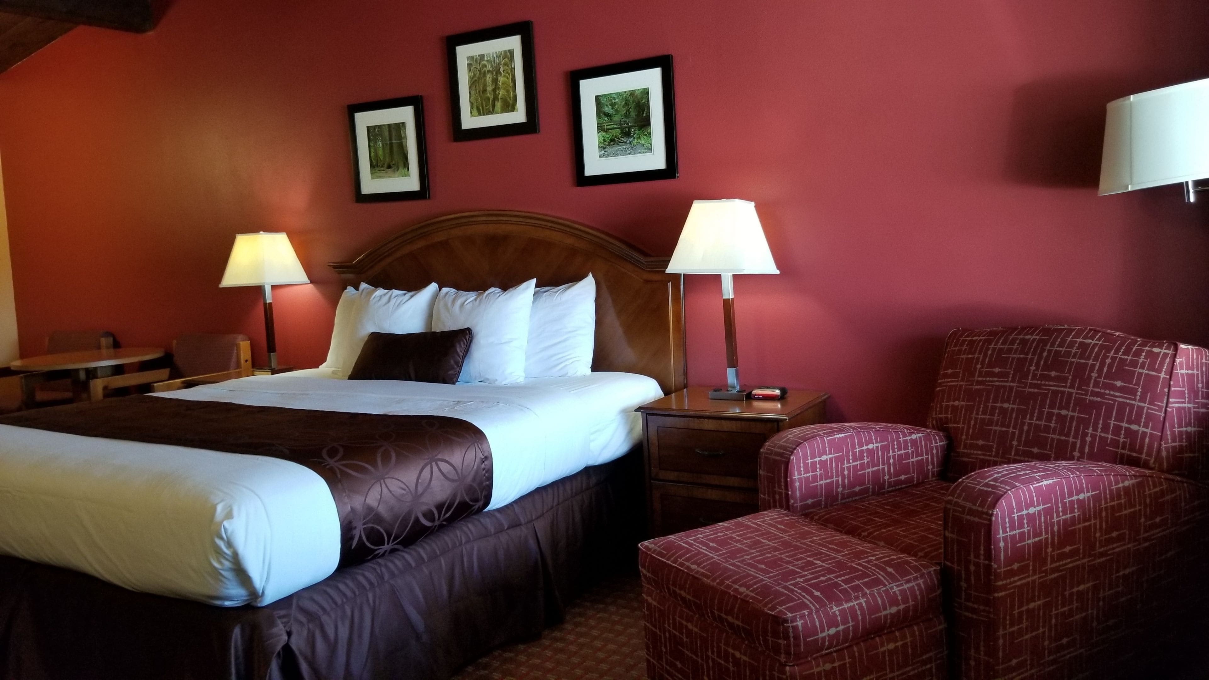 Une chambre d’hôtel avec un grand lit, deux tables de chevet avec des lampes, une tête de lit en bois, un fauteuil rouge et trois tableaux encadrés accrochés au mur.