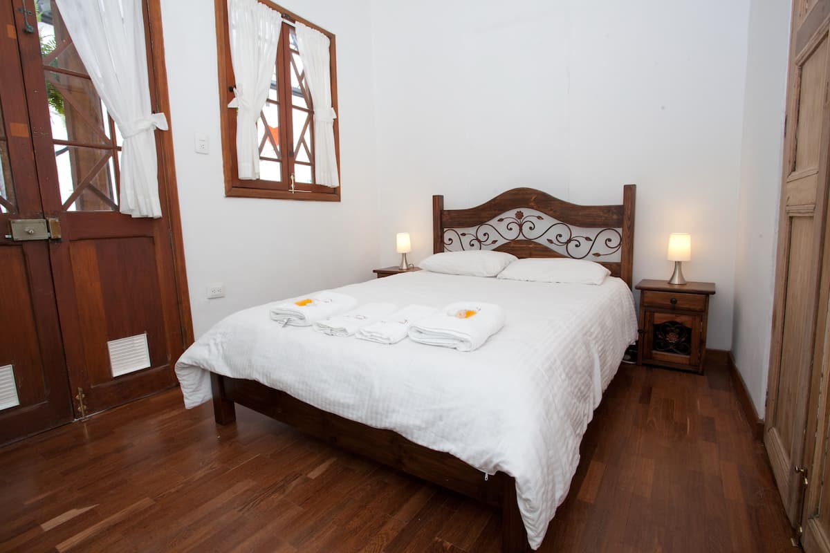 A neatly made bed with white linens and towels, a wooden headboard, two bedside lamps, and a wooden floor.