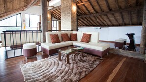 Un vaste espace de vie de style loft, avec un plafond en bois, un canapé d’angle agrémenté de coussins à motifs, une table basse en verre et un tapis à longues mèches.