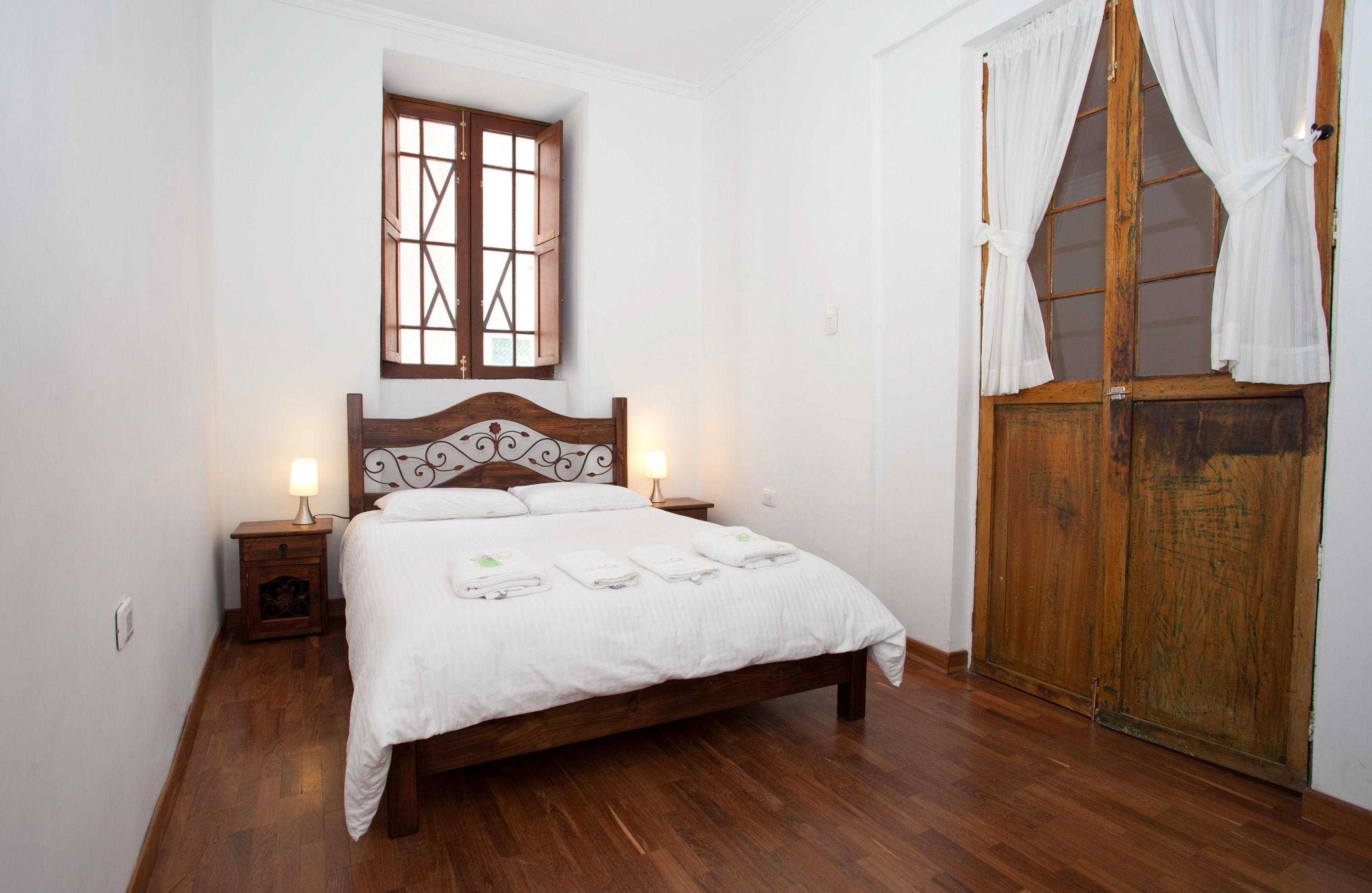 Un dormitorio con una cama de madera, una mesita de noche con una lámpara, una ventana con contraventanas de madera y una puerta de madera con cortinas.