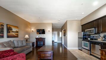 A spacious living room with a sofa, a coffee table, and a TV. There is a kitchen with modern appliances and cabinets.