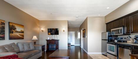 A spacious living room with a sofa, a coffee table, and a TV. There is a kitchen with modern appliances and cabinets.