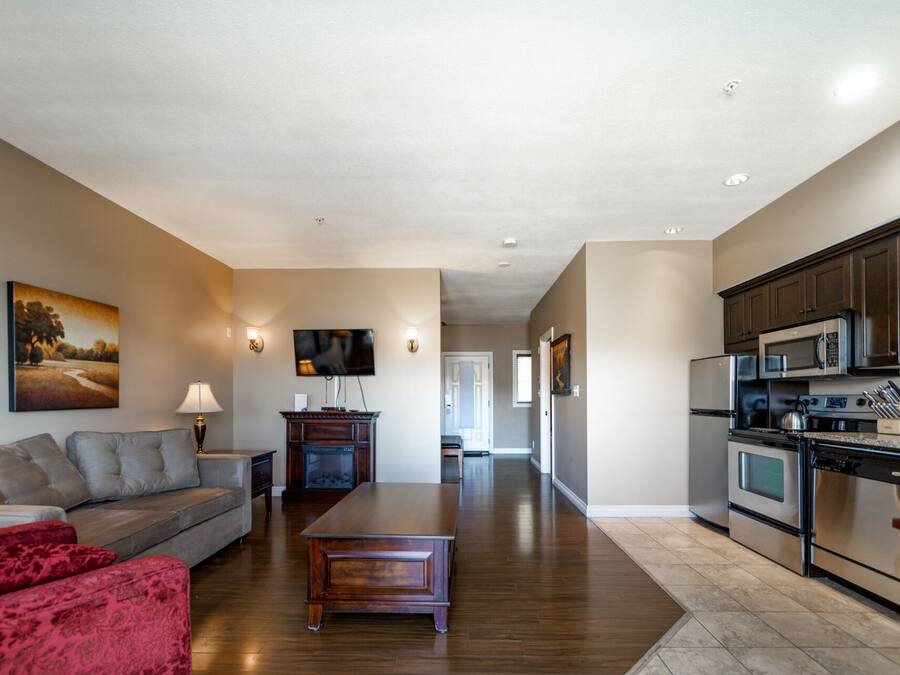 A spacious living room with a sofa, a coffee table, and a TV. There is a kitchen with modern appliances and cabinets.