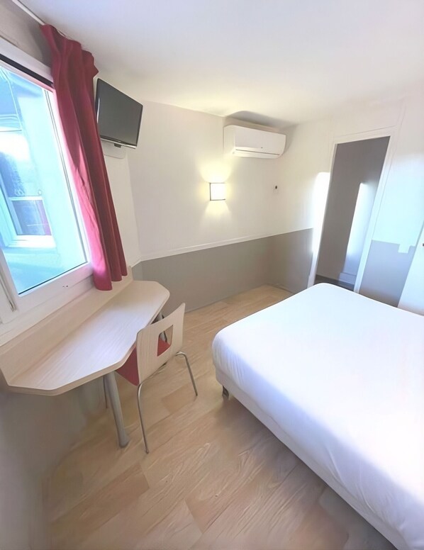 Habitación de hotel con cama, escritorio, silla, televisor montado en la pared y ventana con cortinas.