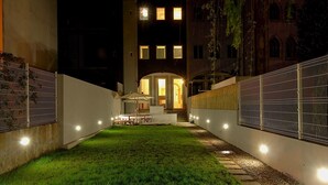 Vista nocturna de un patio con un área de estar al aire libre bien iluminada, una escalera que conduce a la entrada de un edificio y un muro blanco con una cerca de metal.