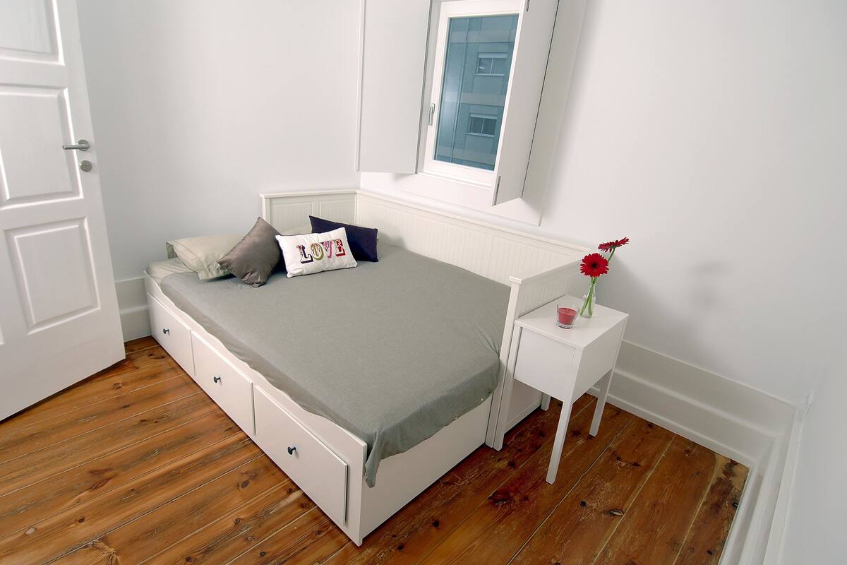 Una cama bien hecha con una colcha gris y almohadas, una mesita de noche blanca con un florero de flores rojas, y una ventana con vista a edificios.