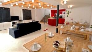 Un espacio habitable moderno con un sofá negro, una mesa de comedor de madera dispuesta para el desayuno y una zona de cocina con armarios rojos.