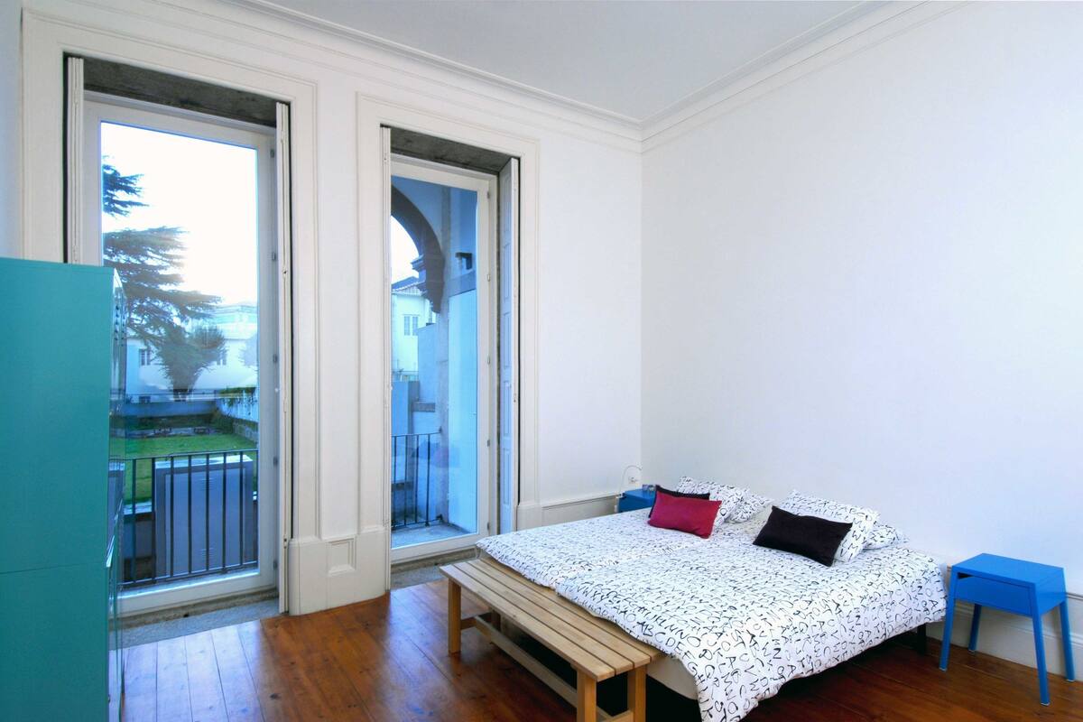 Un dormitorio con cama, un banco, una mesita de noche azul y vistas a través de una puerta.