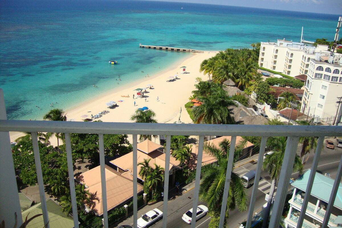 A beach view from a balcony overlooking a sandy beach with clear turquoise water, a pier, and buildings with balconies.