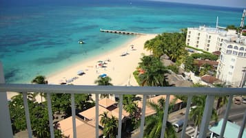 A beach view from a balcony overlooking a sandy beach with clear turquoise water, a pier, and buildings with balconies.