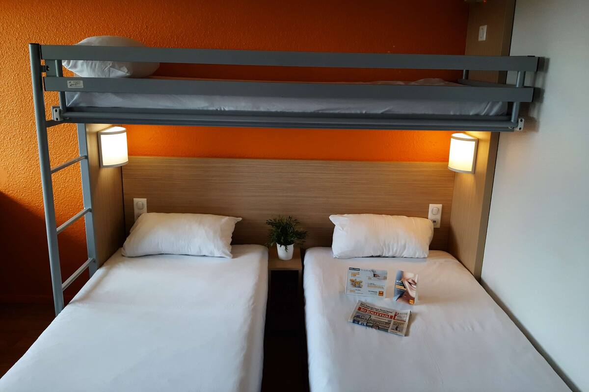 A bunk bed room with two beds, each with a pillow and a bedside lamp. There is a small potted plant between the beds and a magazine on the lower bed.