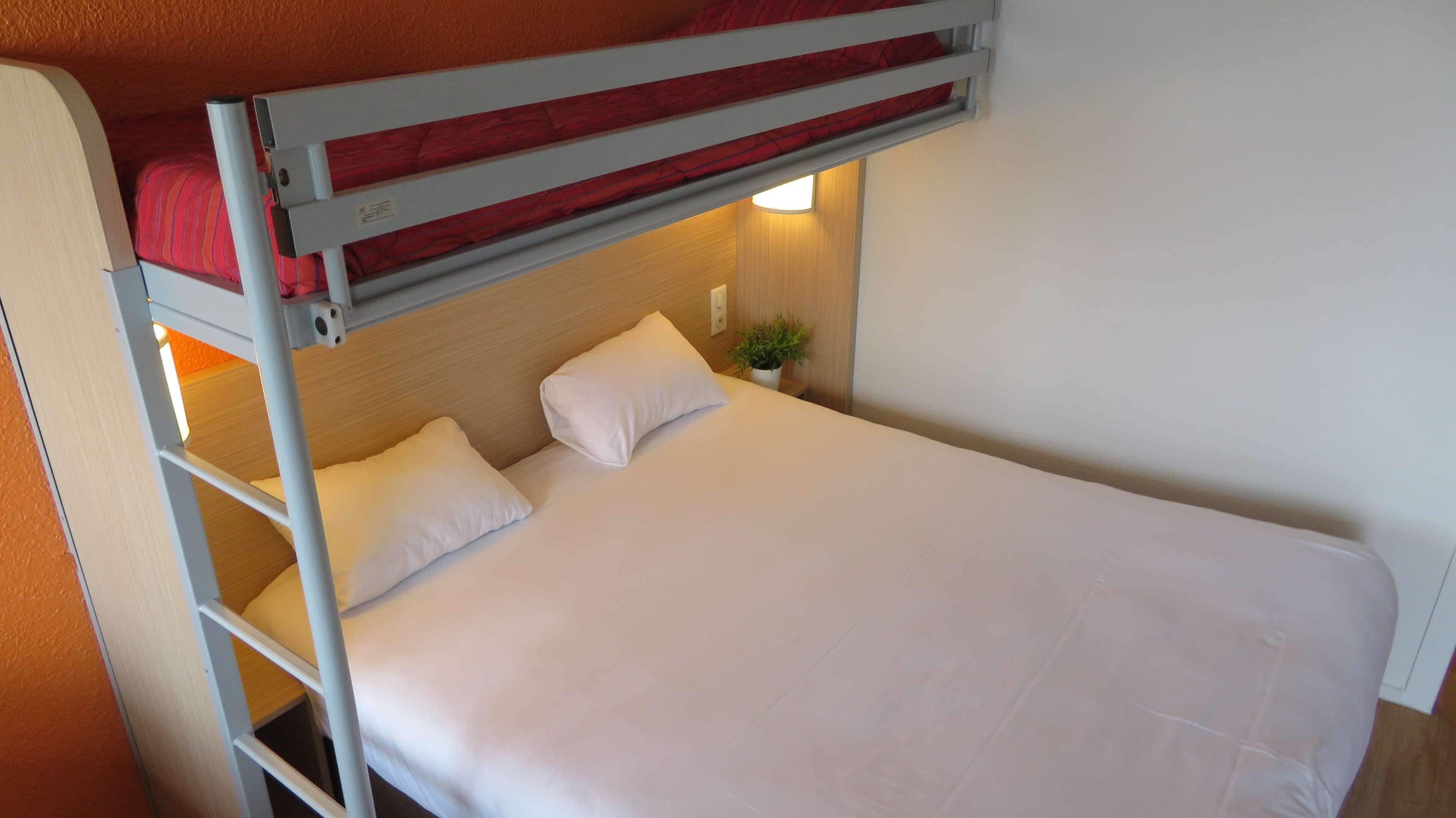 Una cama litera con un colchón rojo en la parte superior y una cama individual abajo. Hay un almohadón blanco en la cama inferior y una pequeña planta en maceta sobre la mesita de noche.