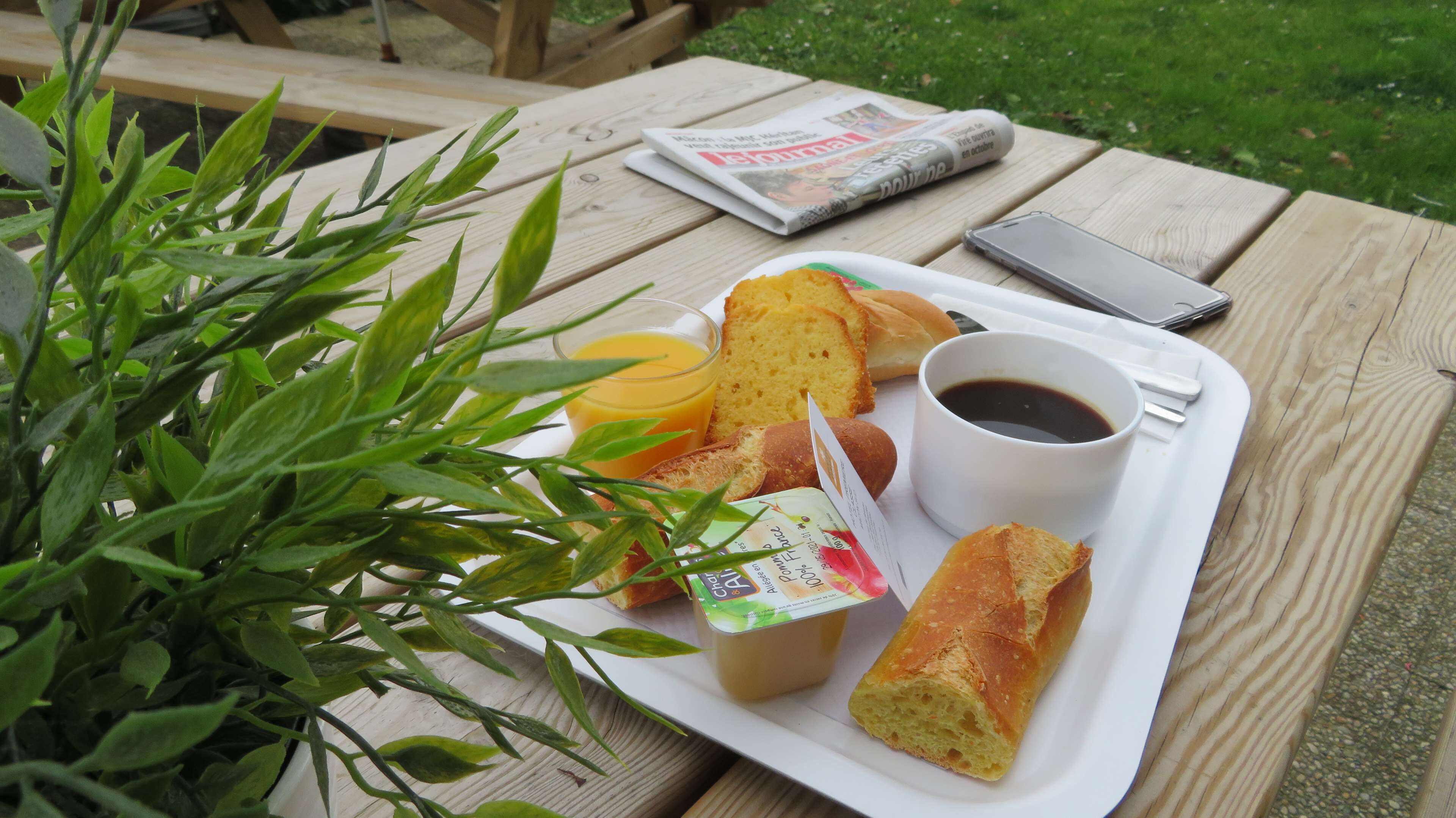 Une table de pique-nique avec un plateau contenant du pain, une boisson et un pot de yaourt, le tout posé sur une table en bois à l’extérieur.