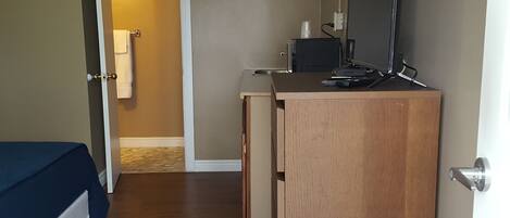 Un dormitorio con cama, cómoda de madera, escritorio con computadora y monitor, y puerta que da a otra habitación.