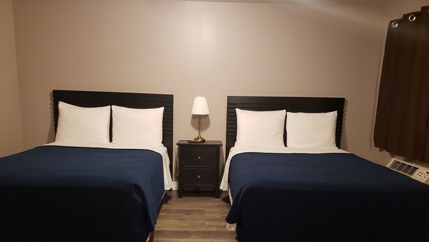 Quarto de hotel com duas camas, cada uma com colcha azul escura e almofadas brancas, uma pequena mesa de cabeceira com abajur, e um aquecedor no chão.