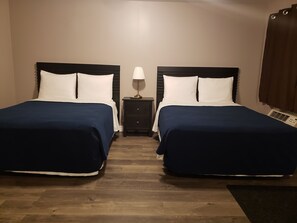Habitación de hotel con dos camas, cada una con cubrecama azul oscuro y almohadas blancas, una mesita de noche con lámpara y un calentador en el suelo.
