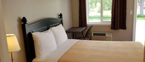 A neatly made bed with white and beige bedding, a dark wooden headboard, a bedside table with a lamp, a window with brown curtains, and a small desk with a chair.