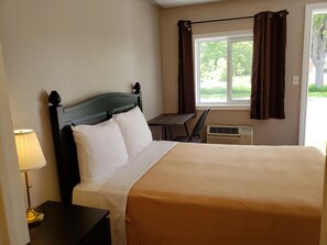 A neatly made bed with white and beige bedding, a dark wooden headboard, a bedside table with a lamp, a window with brown curtains, and a small desk with a chair. - Palace Inn Motel (Sarnia)